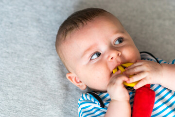 cute caucasian baby boy with blue eyes in striped clothes biting a toy teether, lying down on gray blanket. Teething solution.