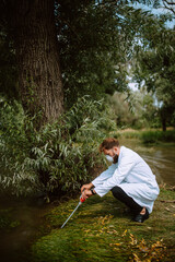 Male caucasian scientist biologist and researcher in protective suit with mask taking water samples from polluted river.