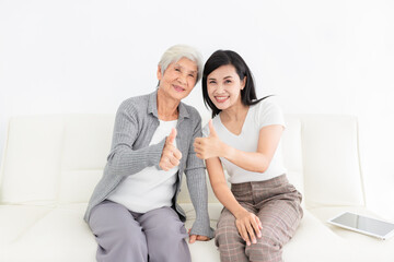 young Asian female and old female show thumbs up sign with hands in home, they feeling happy and smile, they sitting on sofa, mother's day, happiness family time