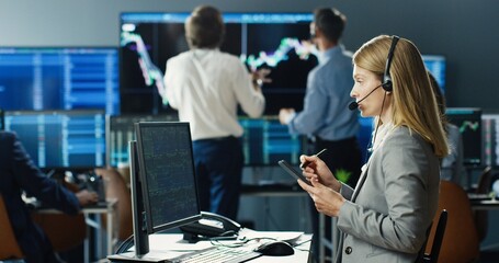Female stock trader working at stock exchange office using digital tablet computer on background of multiple monitors showing data, ticker numbers and graphs.