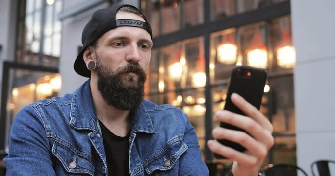 Caucasian handsome young male hipster with piercing and in hat sitting outdoor at table on cafe terrace and having videochat on smartphone. Guy talking and videochatting on mobile phone webcam.