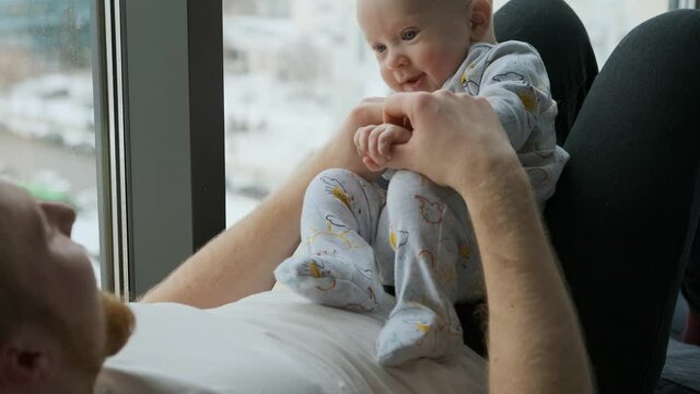 father plays with baby son sitting on his stomach by the window