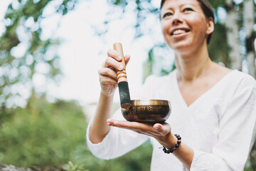 Young smiling woman playing on brass Tibetan singing bowl outdoor. Sound therapy and meditation