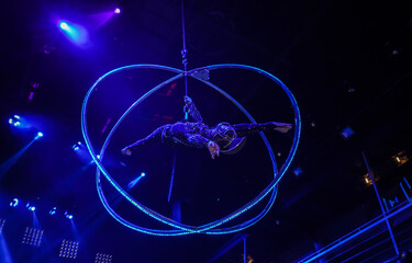 Flexible young woman make performance on aerial hoop, flexible back on aerial hoop, aerial circus show, blue light. Flexible woman gymnast upside down on hoop. Night club performance