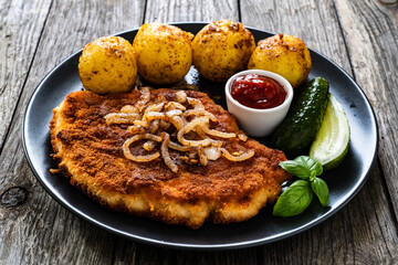 Breaded pork chop with boiled potatoes and cucumber served on black plate on wooden table