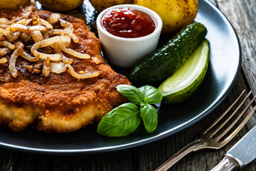 Breaded pork chop with boiled potatoes and cucumber served on black plate on wooden table