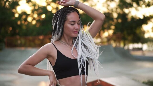 Sensual woman in black top with black and white dreadlocks dancing in empty skatepark. Girl relaxfully dancing with closed eyes. Wearing silver neckless. Trees in sunlight on the background