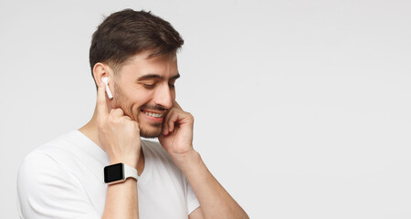 Banner of young man in white t-shirt, wearing smartwatch and listening to music via wireless earphones, isolated on gray background