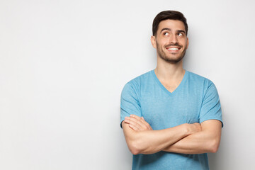 Fototapeta premium Portrait of young handsome guy wearing blue T-shirt standing with crossed arms, looking up and smiling with curious face, isolated on gray background with copy space