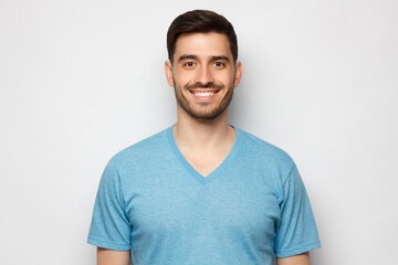 Close-up portrait of young smiling handsome male with short dark hair and stubble, wearing blue T-shirt, isolated on gray background