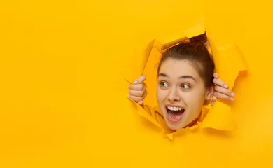 Foto op Aluminium Excited girl looking through hole in paper at left with eyes round with surprise, isolated on yellow background with copy space © Damir Khabirov