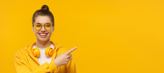 Banner of young laughing girl wearing headphones around neck, pointing right, isolated on yellow...