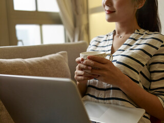 young asian woman working at home sitting on couch enjoying a cup of coffee