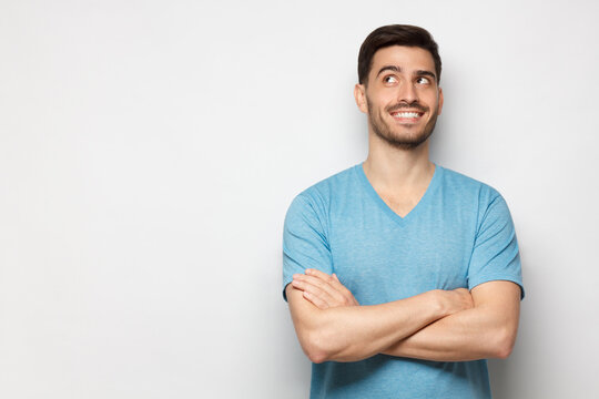 Portrait of young handsome guy wearing blue T-shirt standing with crossed arms, looking up and smiling with curious face, isolated on gray background with copy space - Powered by Adobe