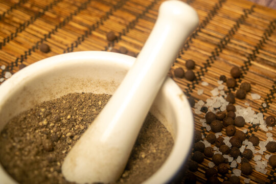 Ground black allspice in a ceramic mortar and black allspice and coarse salt scattered on a reed Mat. Close up.