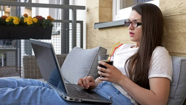 Young attractive woman is sitting comfortably on the balcony of her apartment with a laptop.