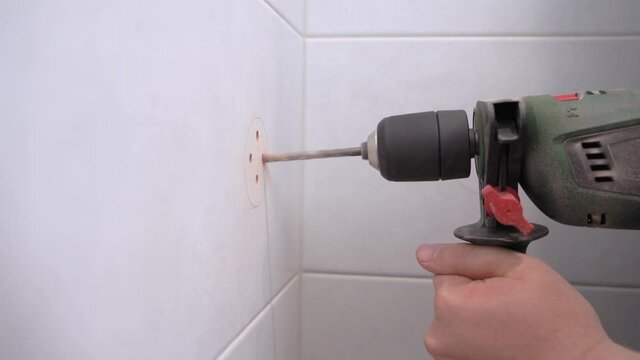 Husband or handyman uses electric drill to make holes according to plan in surface of tile on the wall to hang corner shelf or hook for towels in bathroom, side view.
