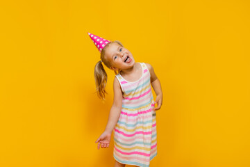 Happy birthday child girl with two ponytales in pink cap on colored yellow background showing her tongue.