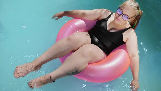 Top view of young overweight girl dressed in swimsuit swims in outdoor pool on pink inflatable swimming ring. Body positivity
