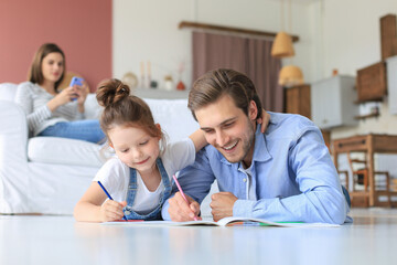 Fototapeta premium Happy father smilling daughter lying on warm floor enjoying creative activity, drawing pencils coloring pictures in albums, mother resting on couch, family spend free time together.