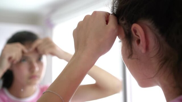 teenage girl with long hair in ponytail pops zits reflecting in bathroom mirror at home in early morning close view