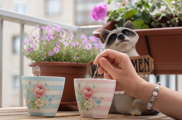 Girl with jewelry, rings and bracelets on her hand drinking a lovely cup of cappuccino coffee from a nice porcelain cup with a small cute raccoon sculpture in the background with a sign Welcome
