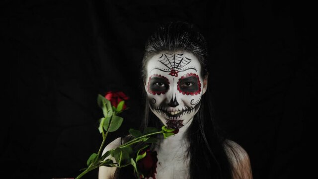 Halloween mask Calavera Catrina. Girl with a rose in her hands. Mexican day of the dead. Portrait of a young woman with a terrible multi-colored makeup for Halloween on a dark background. 4K. 