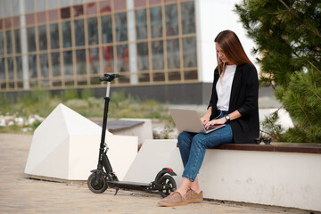 Young contemporary yoman with laptop sitting by green tree in urban environment