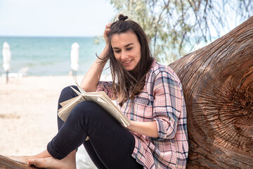 A happy girl reads a book on a tree, against the background of a sea beach. The person abstracted from everything.