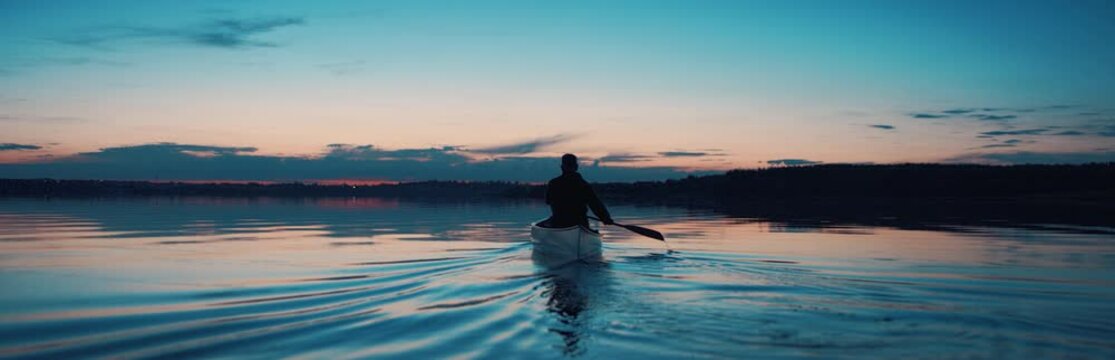 WIDE Man canoeing in a traditional wooden boat on a large lake at dawn. Shot on RED cinema camera with 2x anamorphic lens
