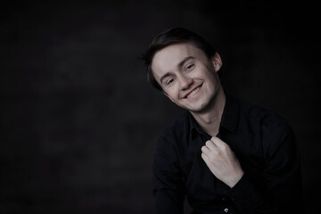 Fototapeta premium Happy young man. Close-up Portrait of handsome young smiling man in casual black shirt touching his collar and smiling while standing against black background. Lauphing handsome boy.