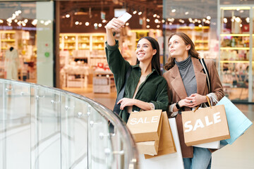 Young cheerful woman with smartphone and paperbags making selfie with friend