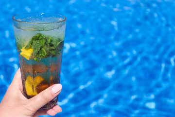 Mojito cocktail with fresh mint and lemon on a background of blue water of the pool. Ice drinks, summer concept. Close-up, copy space for text