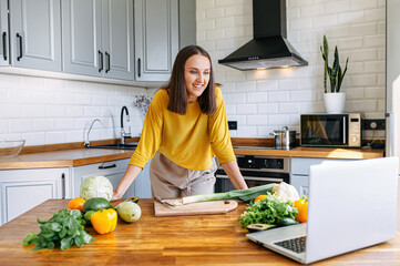 A young woman in yellow shirt cooks in the kitchen and looks with interest at the laptop screen. Woman prepare veggie dish, vegetables on the table