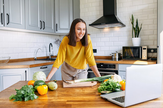 A Young Woman In Yellow Shirt Cooks In The Kitchen And Looks With Interest At The Laptop Screen. Woman Prepare Veggie Dish, Vegetables On The Table