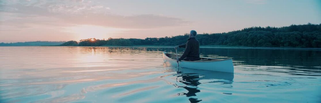 Man canoeing in a traditional wooden boat on a large lake at dawn, enjoying sunrise. Shot on RED cinema camera with 2x anamorphic lens