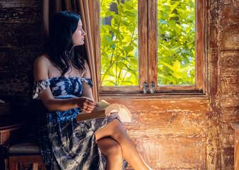 Beautiful young woman sitting by the window in wooden bunglow rustic house and reading a book. Summer exotic vacation