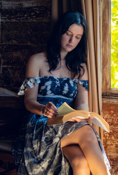 Beautiful young woman sitting by the window in wooden bunglow rustic house and reading a book. Summer exotic vacation