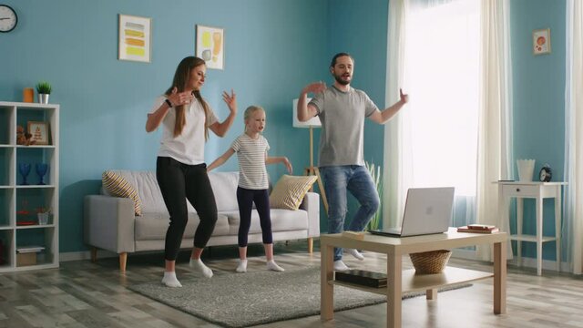 Young family, mother, father, and small girl, are dancing together synchronously, learn new dance, using laptop, repeat movements after online coach, have fun, Slow motion.