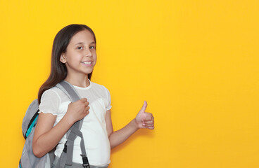 a cute smiling brunette girl schoolgirl with a backpack on her shoulder with bent arms at the elbows shows cool, cool, yes on a yellow background moke up