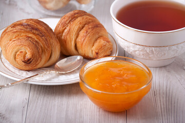 Croissants on a white plate, a vase with apricot jam and a cup with tea