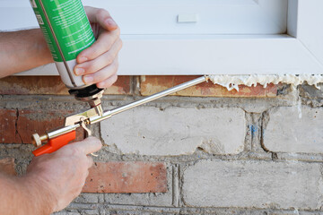 Handyman using polyurethane foam for installing a plastic window. Side view on a worker in action.