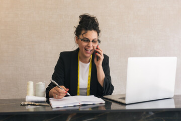 brunette woman with curly, upturned hair and round glasses smiling and talking on a mobile phone and writing on paper with a grey pen and a laptop next to her 