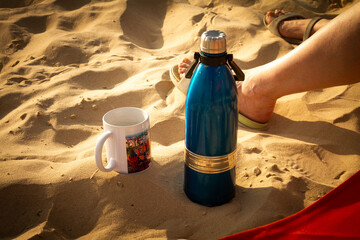 Thermos and a mug are standing on the sea sand in the sunset. A girl sits on a beach chair in...