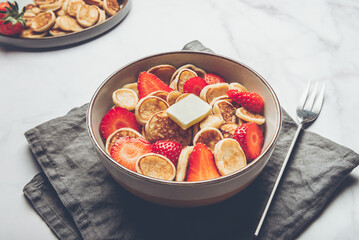 Trendy home breakfast with tiny cereal pancakes with piece of butter and fresh strawberries in the bowl and grey napkin