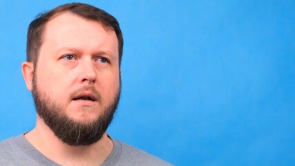 Portrait of a sad bearded man in a gray t-shirt singing and talking to the camera on a blue background.