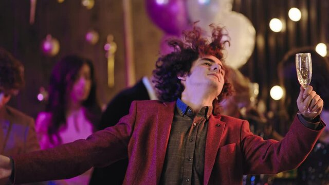 Very charismatic guy with curly hair in front of the camera dancing moving funny enjoying the evening at big glamorous party he holding a glass of champagne. 4k