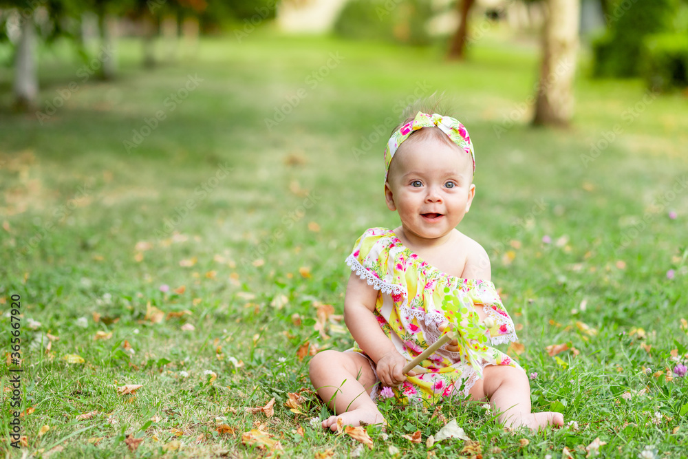 Wall mural little baby girl 7 months old sitting on a green lawn in a yellow dress and playing with a toy, walk