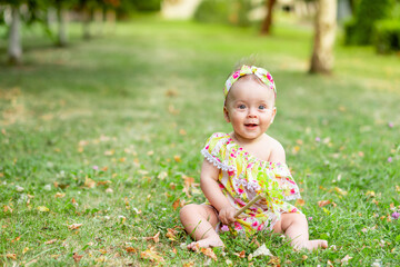 little baby girl 7 months old sitting on a green lawn in a yellow dress and playing with a toy, walking in the fresh air, space for text