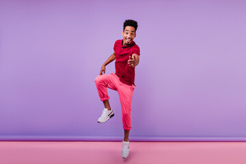 Good-humoured young man pointing finger to camera with cheerful smile. Indoor photo of carefree curly african guy expressing good emotions.